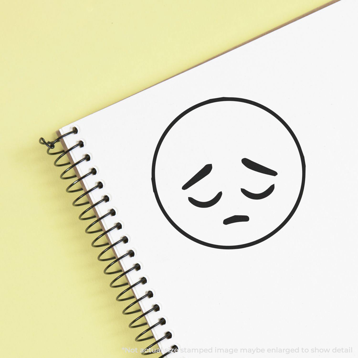 Round Sad Smiley Rubber Stamp impression on a spiral notebook, showing a sad face with downturned eyes and mouth.