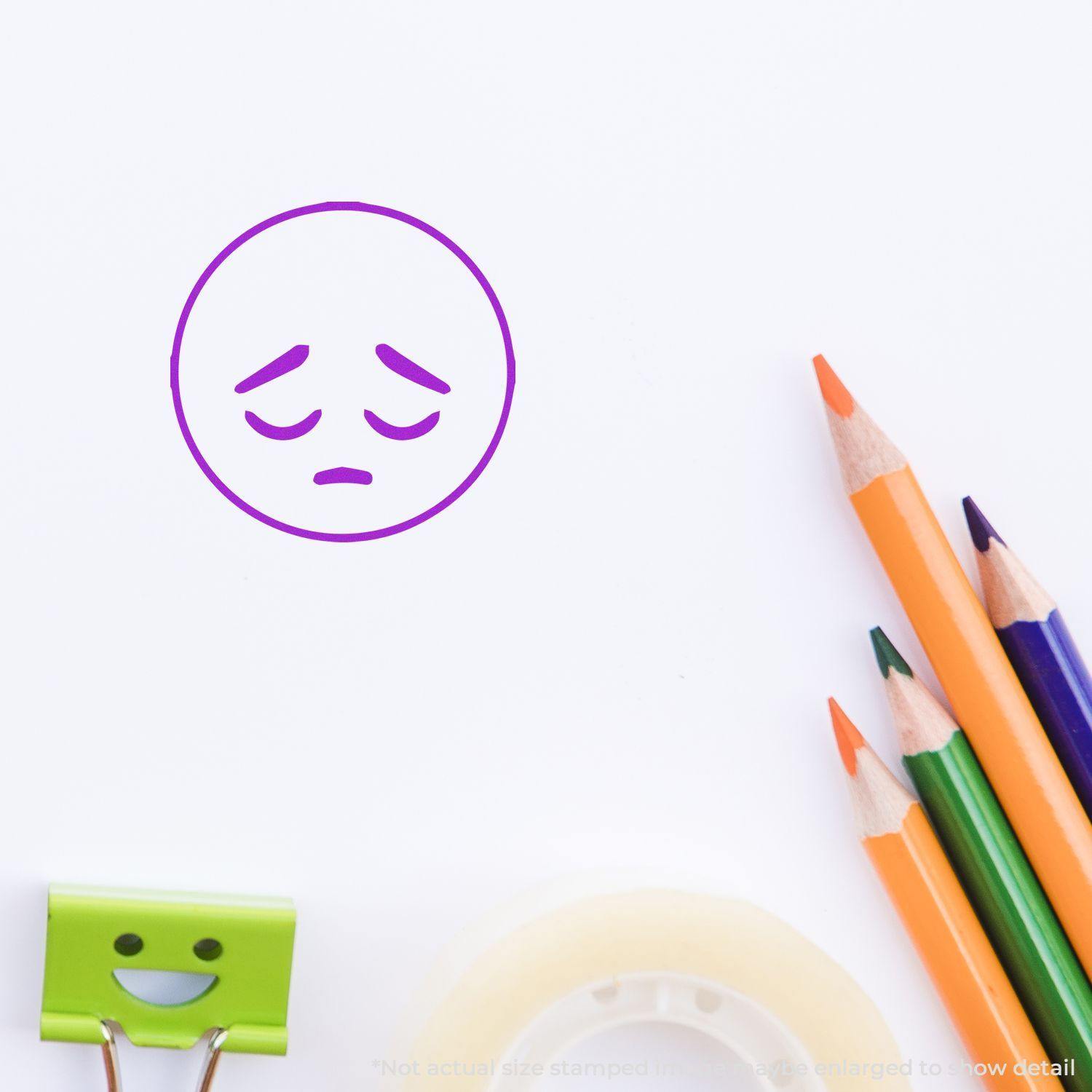 Round Sad Smiley Rubber Stamp in purple ink on white paper, surrounded by colored pencils, a green binder clip, and tape.