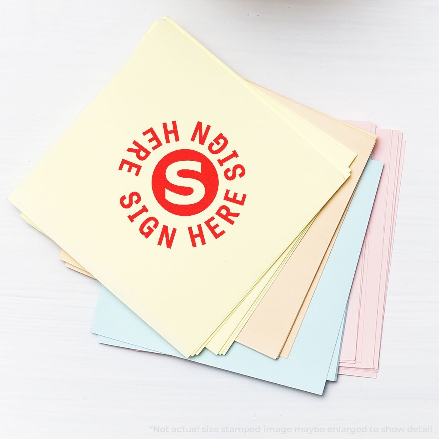 A stack of pastel-colored papers stamped with the Self Inking Round Sign Here Sign Here Stamp in red ink on a white surface.