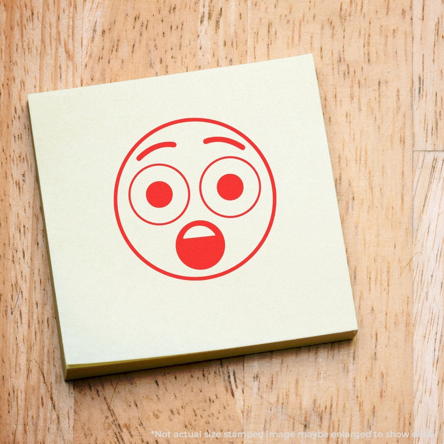 Self Inking Round Surprised Smiley Stamp in red ink on a yellow sticky note, placed on a wooden surface.