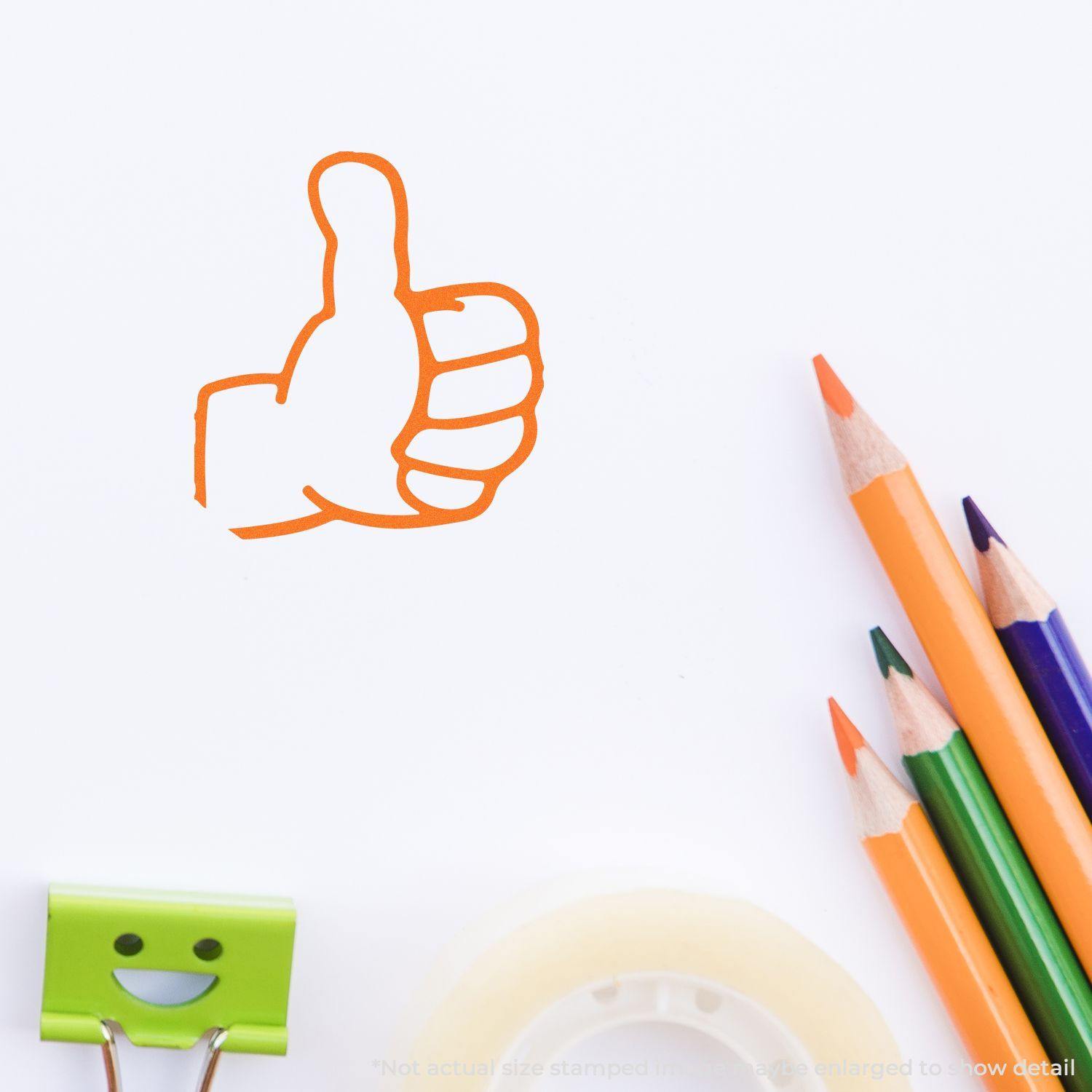 Round Thumbs Up Rubber Stamp in orange ink on white paper, surrounded by colored pencils, a smiley face clip, and tape.