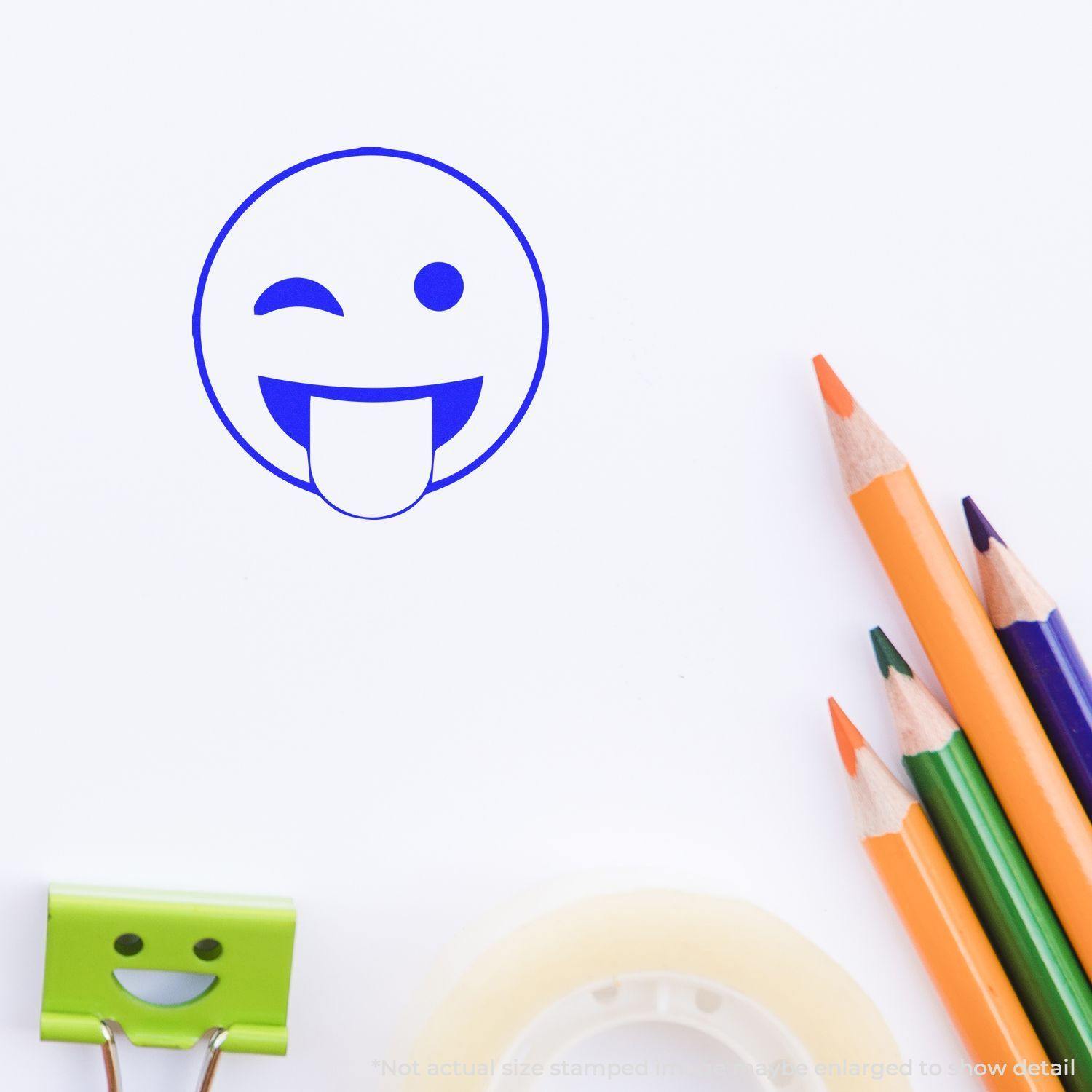 Round Tongue Out Smiley Rubber Stamp in blue ink on white paper, surrounded by colored pencils, a binder clip, and tape.