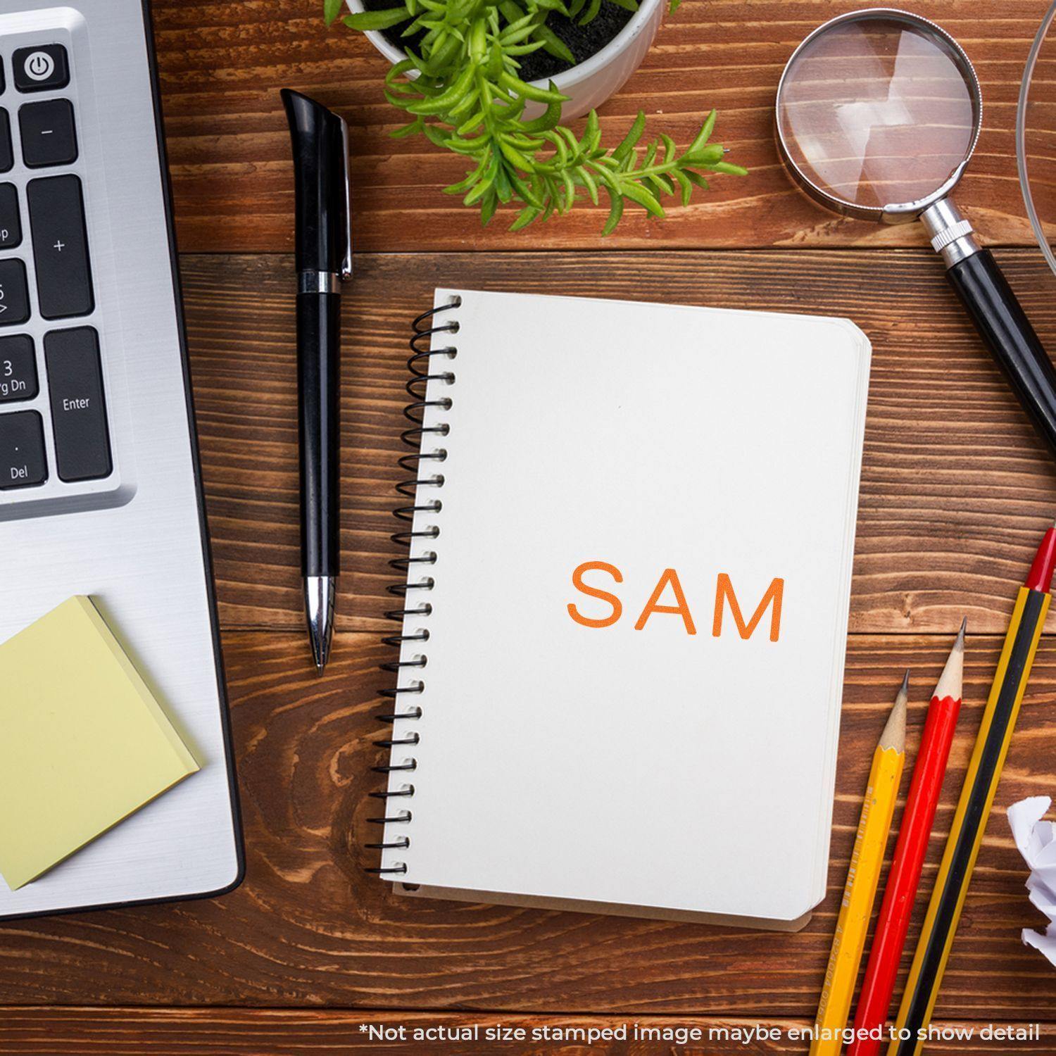 Slim Pre-Inked SAM Stamp on a notebook, surrounded by a laptop, pen, sticky notes, magnifying glass, plant, and colored pencils.