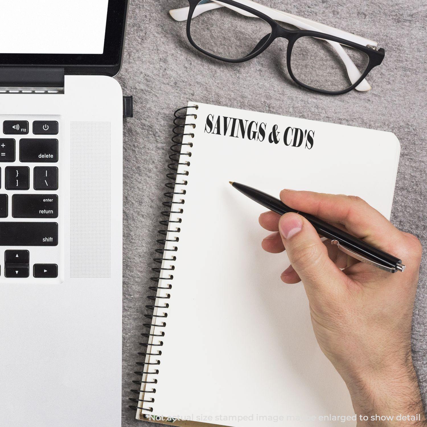 Hand holding a pen next to a Large Self Inking Savings & CD's Stamp on a spiral notebook, with a laptop and glasses nearby.