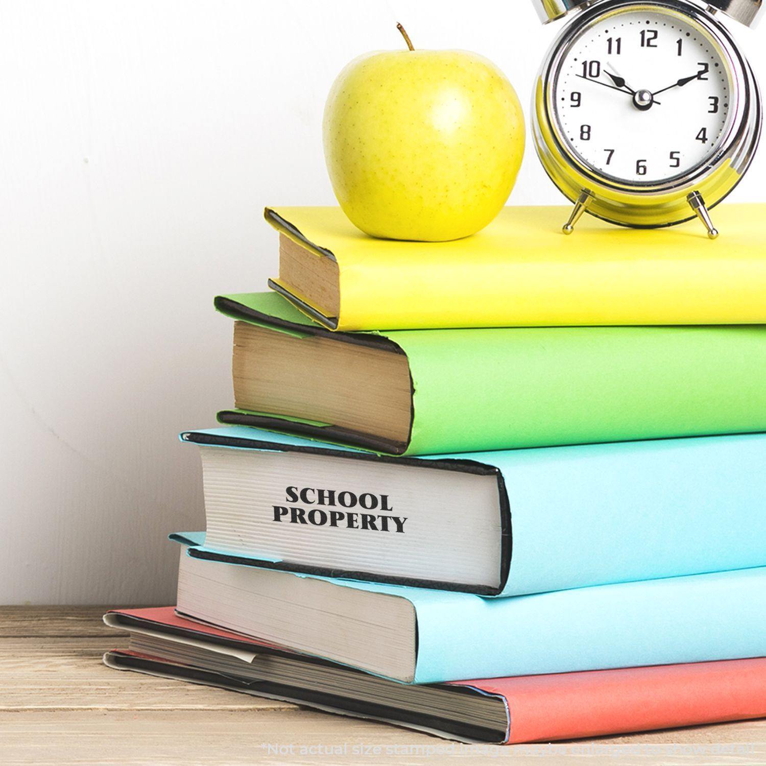 A stack of colorful books with SCHOOL PROPERTY stamped on one, next to an apple and a clock. Large Self Inking School Property Stamp in use.