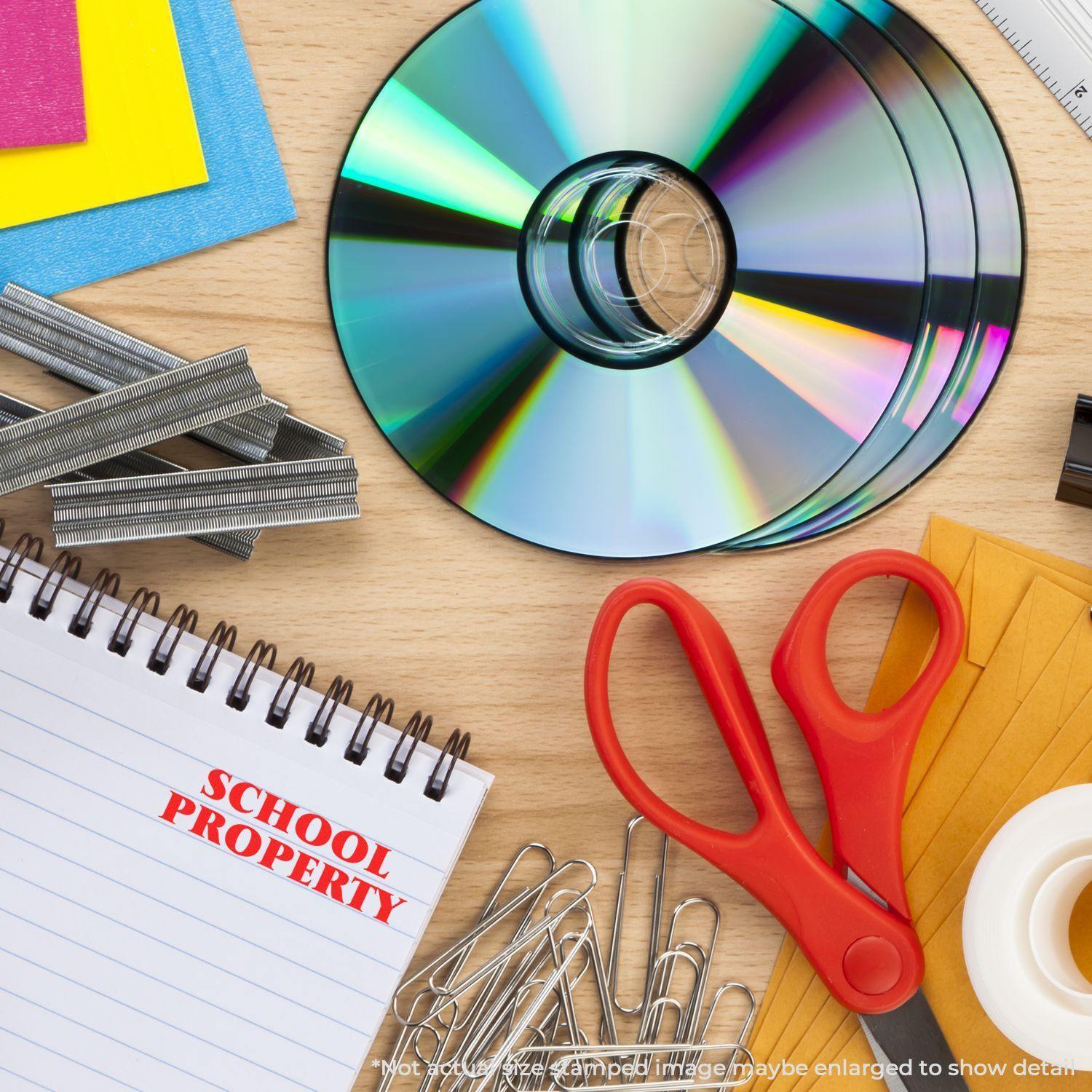 Large Self Inking School Property Stamp marking a notebook, surrounded by CDs, paper clips, red scissors, and colorful paper on a desk.