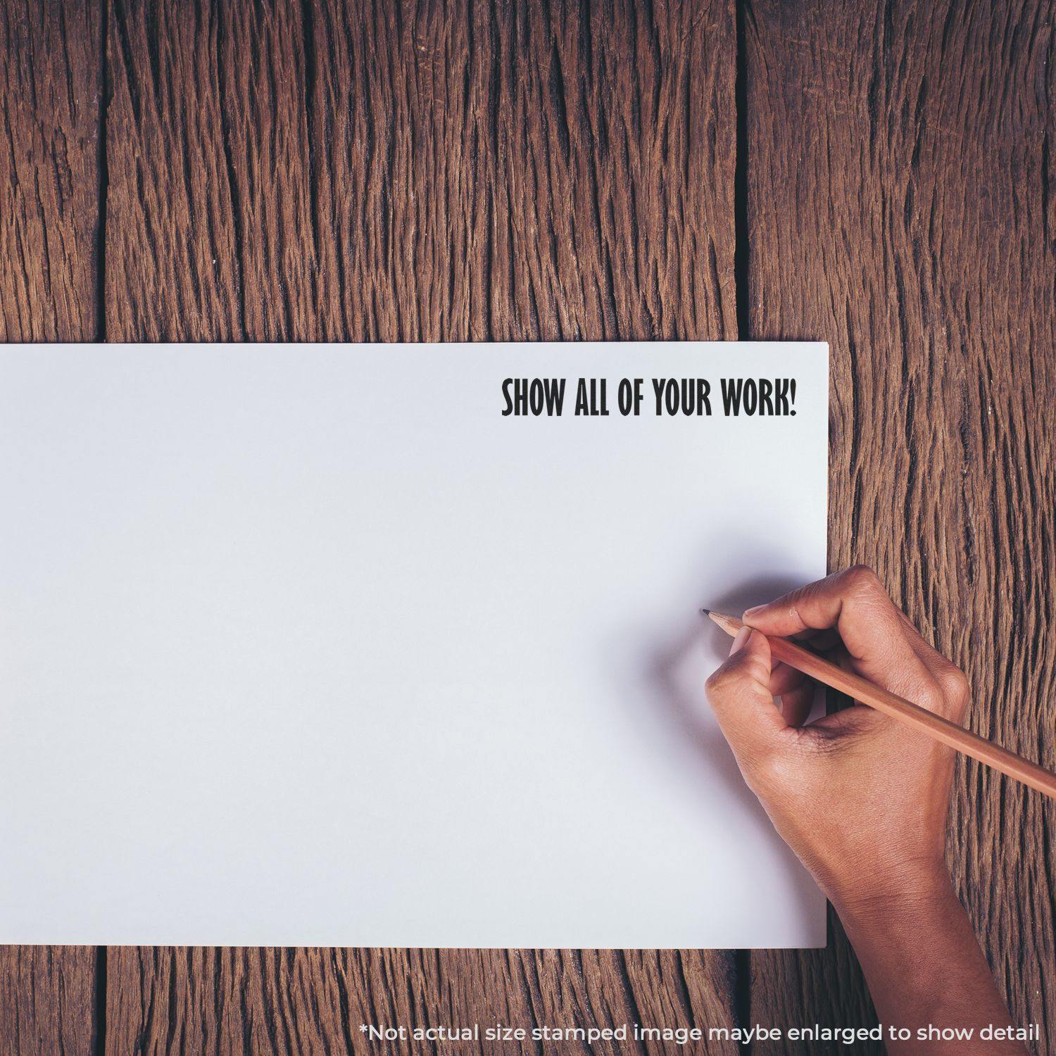 Hand holding pencil near paper stamped with SHOW ALL OF YOUR WORK! using the Large Self Inking Show All Of Your Work Stamp on a wooden surface.