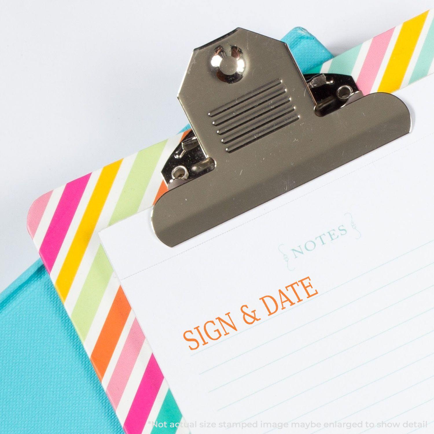 Clipboard with colorful stripes holding a notepad stamped with Sign & Date in orange ink, emphasizing the Sign & Date Rubber Stamp.