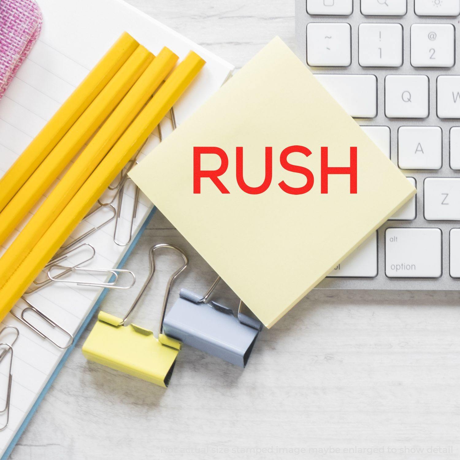 Large Pre-Inked Skinny Rush Stamp in use, stamping RUSH on a yellow sticky note, surrounded by pencils, paper clips, and a keyboard.