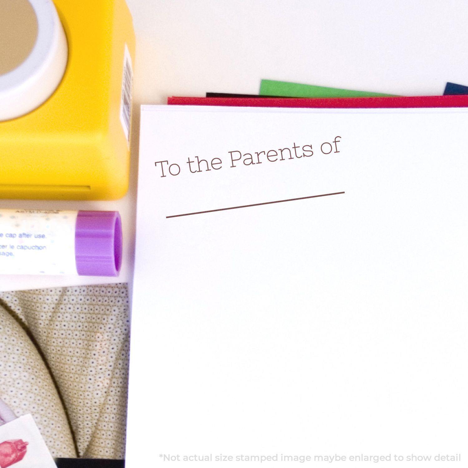 Large Self Inking Skinny To the Parents of Stamp in use on a white paper, surrounded by stationery items like glue stick and colored paper.