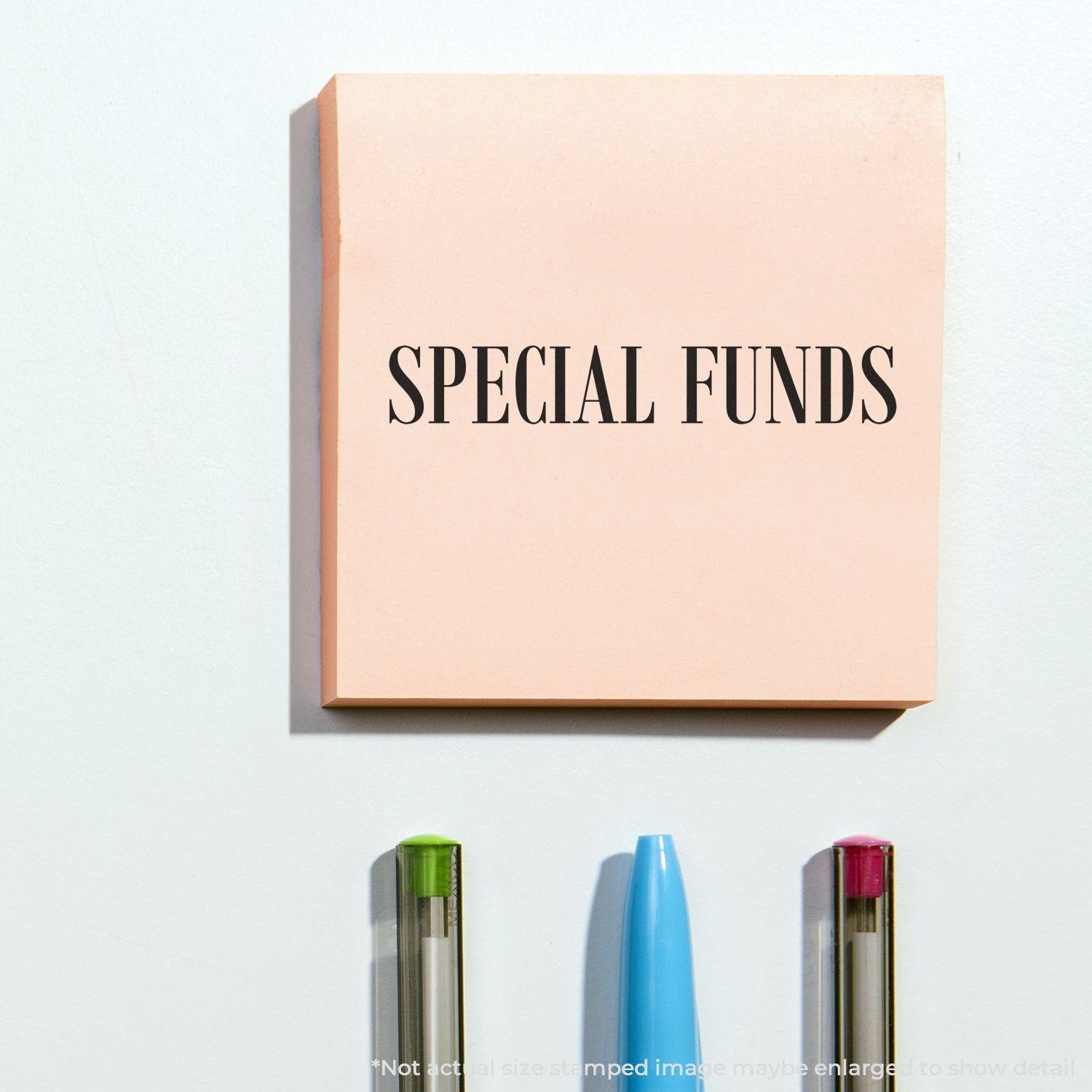 Large Self Inking Special Funds Stamp used on a pink sticky note, with three pens below it on a white surface.