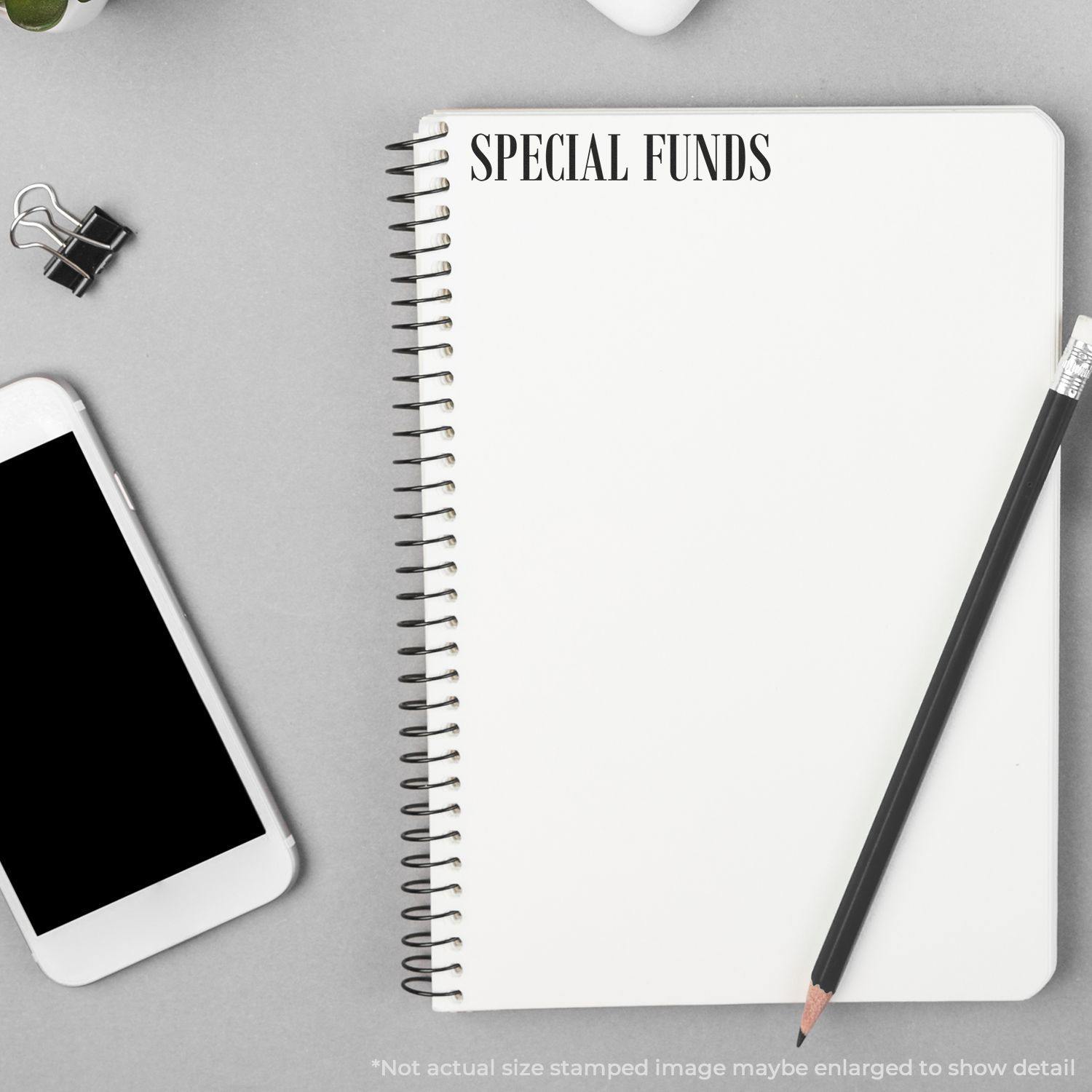 Self Inking Special Funds Stamp used on a spiral notebook, placed on a desk with a smartphone, pencil, and binder clip.