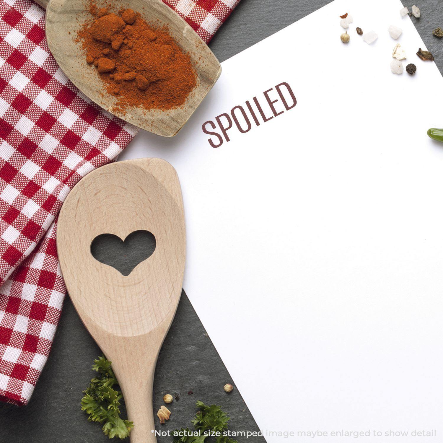 Self Inking Spoiled Stamp marking SPOILED on paper, with a wooden spoon, spices, and a red checkered cloth in the background.