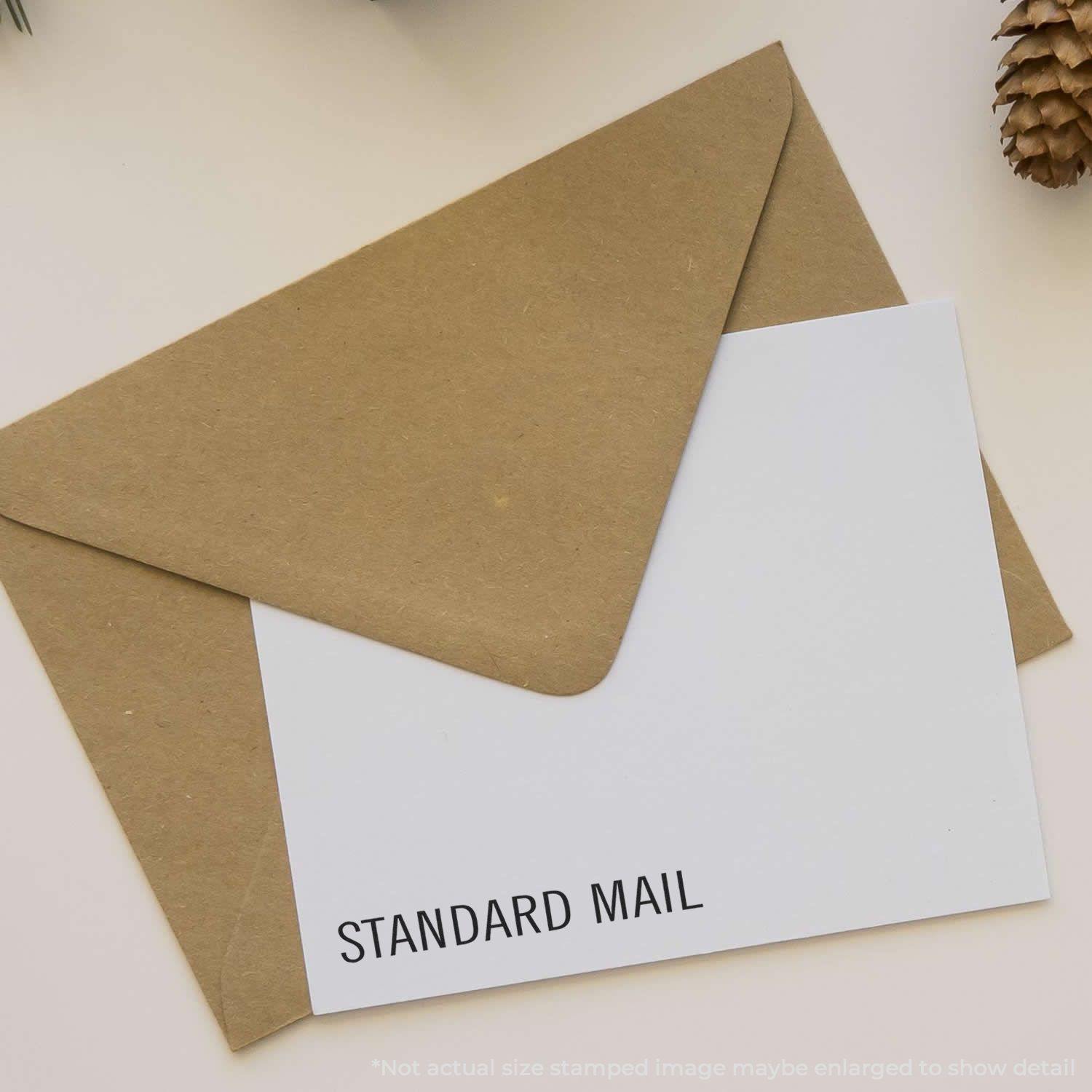 Envelope stamped with STANDARD MAIL using the Large Self Inking Standard Mail Stamp, placed on a beige surface with a pinecone nearby.