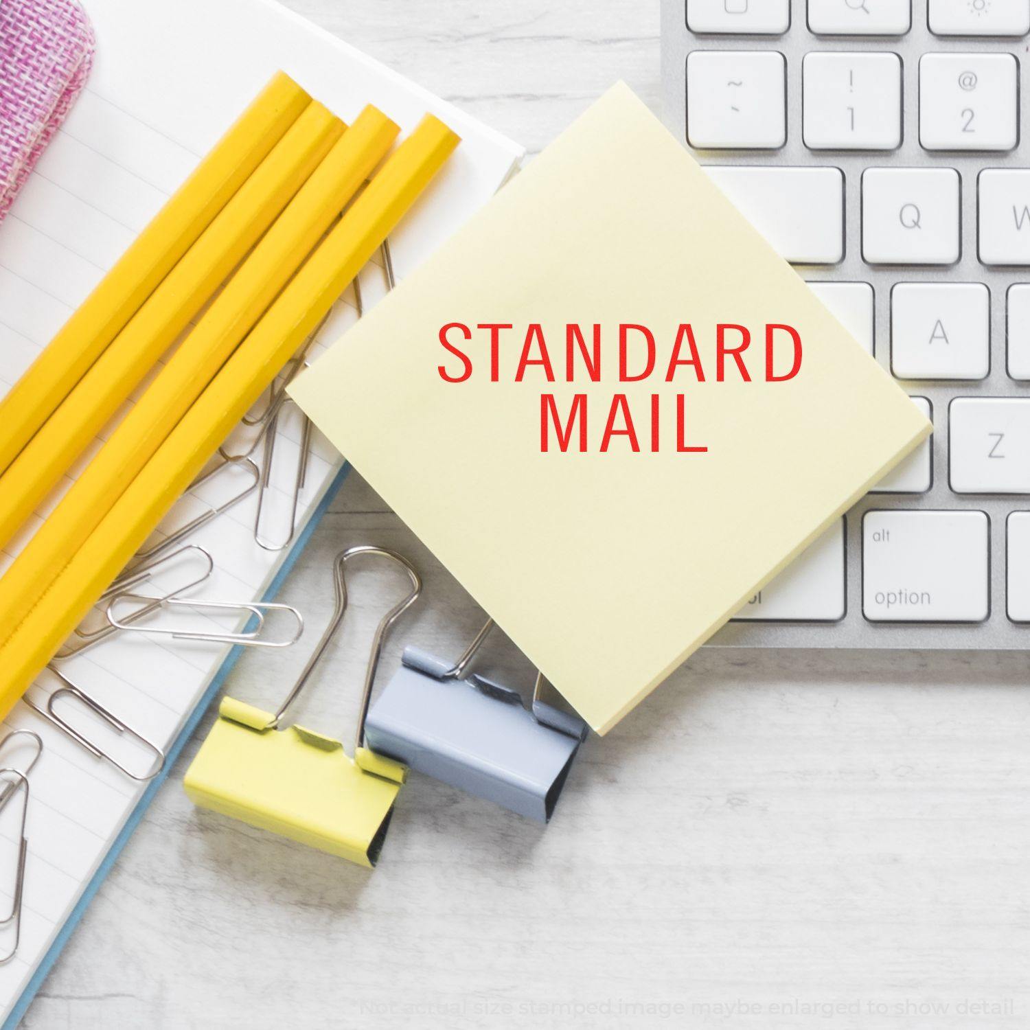 Large Pre-Inked Standard Mail Stacked Stamp on a yellow sticky note, next to a keyboard, pencils, and paper clips.