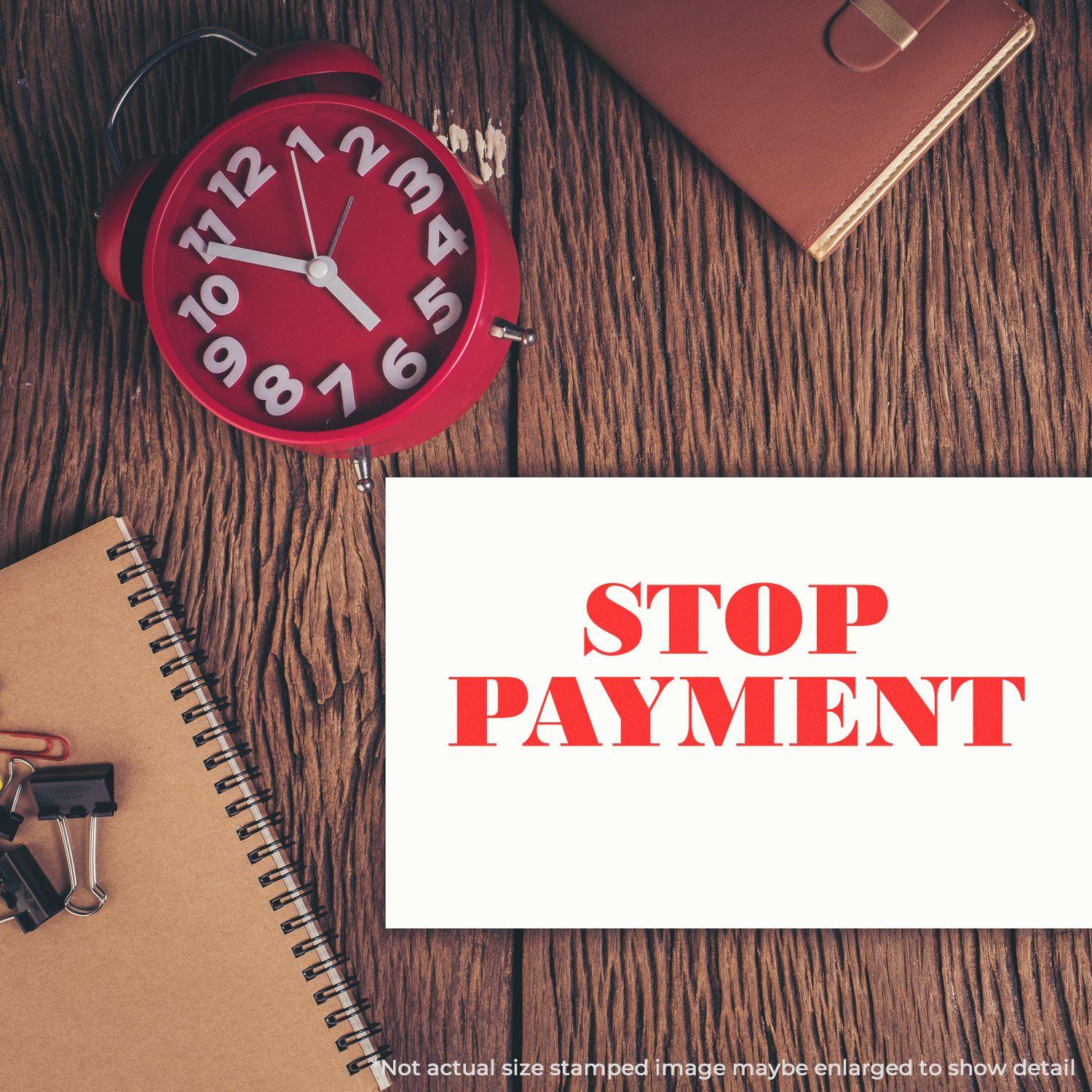 Self Inking Stop Payment Stamp on white paper, surrounded by a red clock, notebook, and office supplies on a wooden desk.