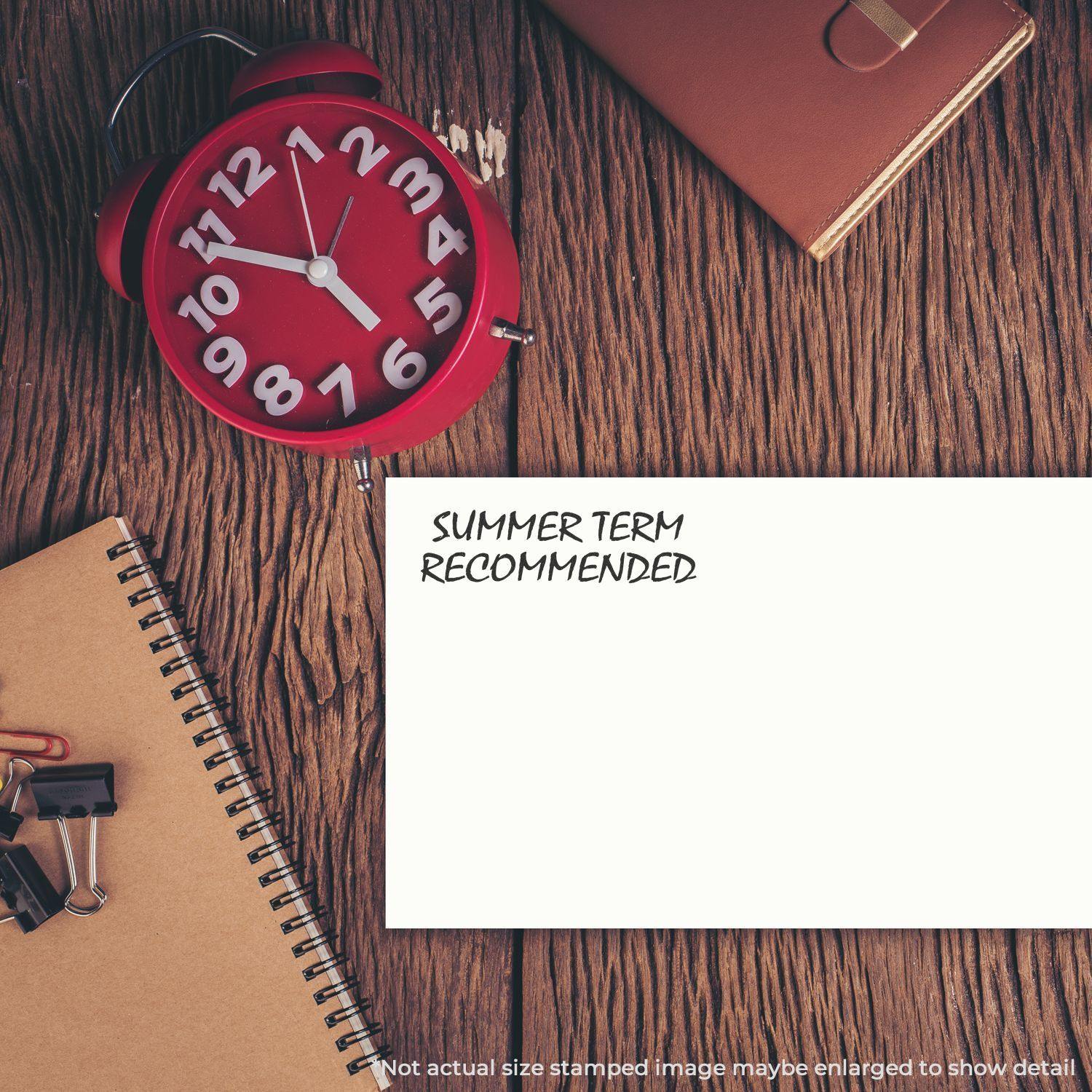 Large Pre-Inked Summer Term Recommended Stamp on a white paper, surrounded by a red clock, notebooks, and office supplies on a wooden desk.