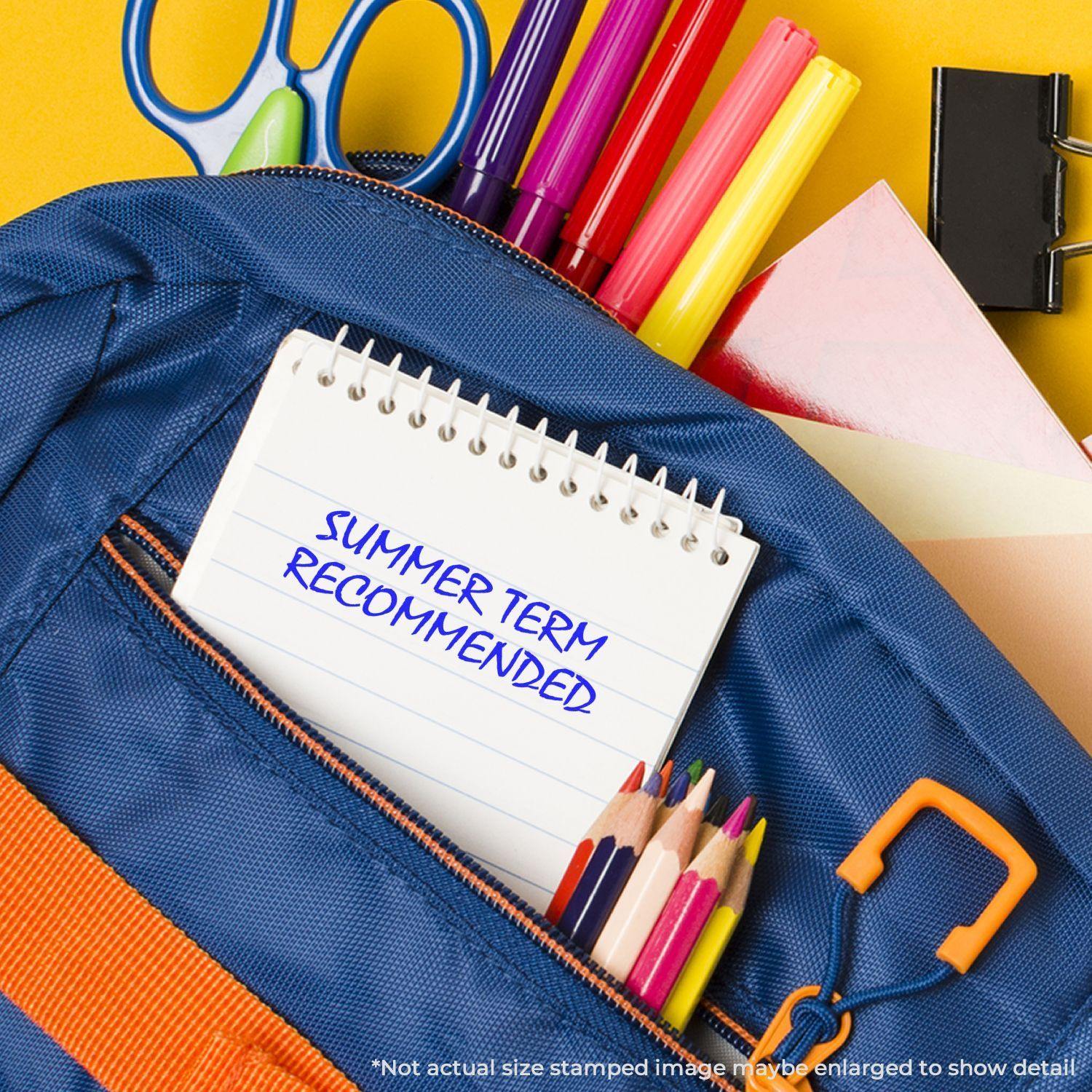 A Slim Pre-Inked Summer Term Recommended Stamp is used on a notepad placed in a blue backpack with school supplies.