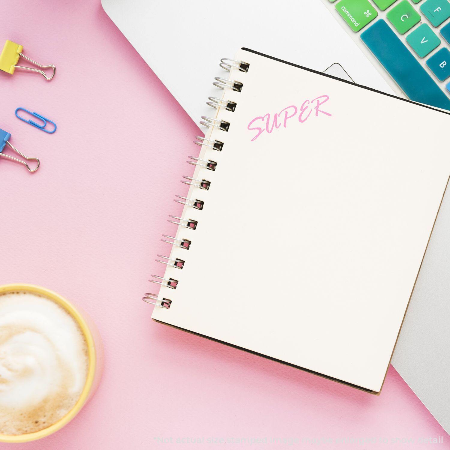 A Slim Pre-Inked Super Stamp is used to stamp the word SUPER in pink on a spiral notebook, placed on a pink desk with a coffee cup.