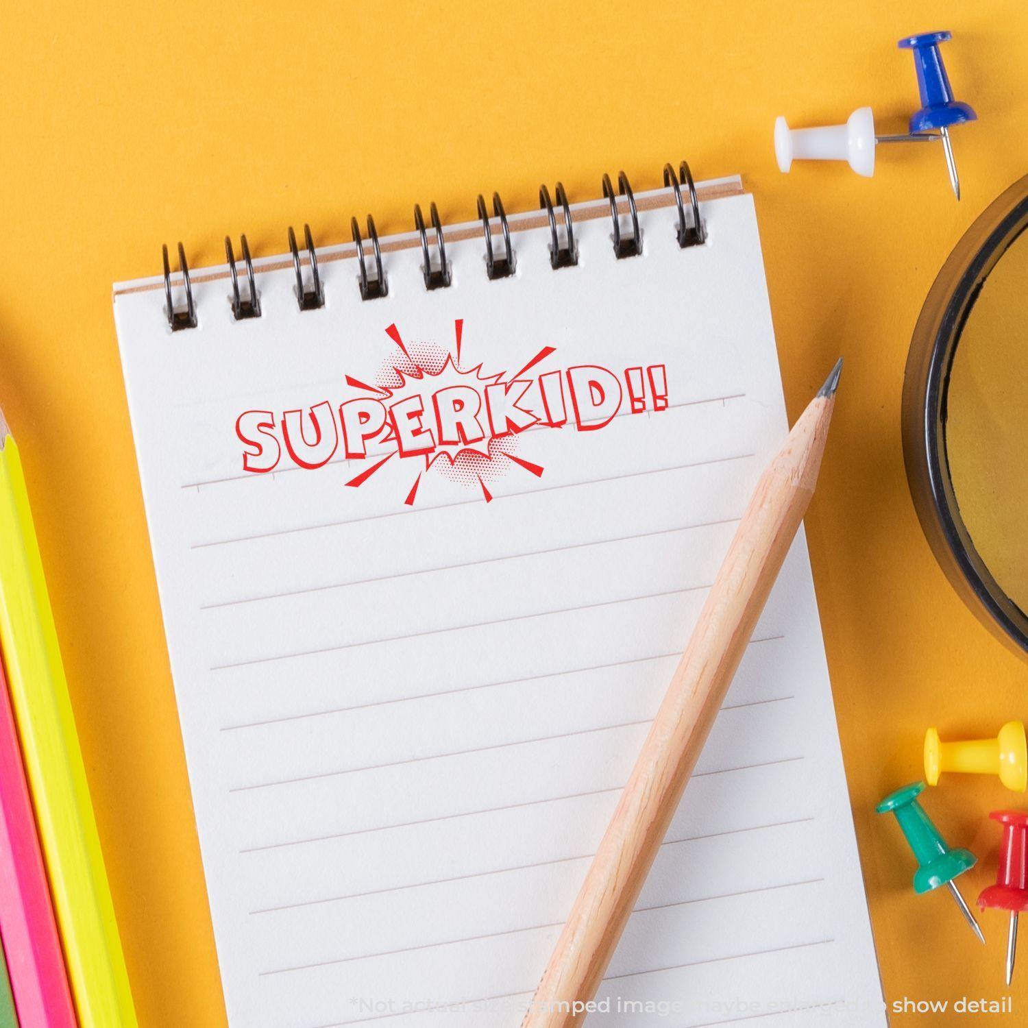 Slim Pre-Inked Superkid Stamp impression on a notepad, surrounded by colorful pencils, push pins, and a pencil on a yellow background.