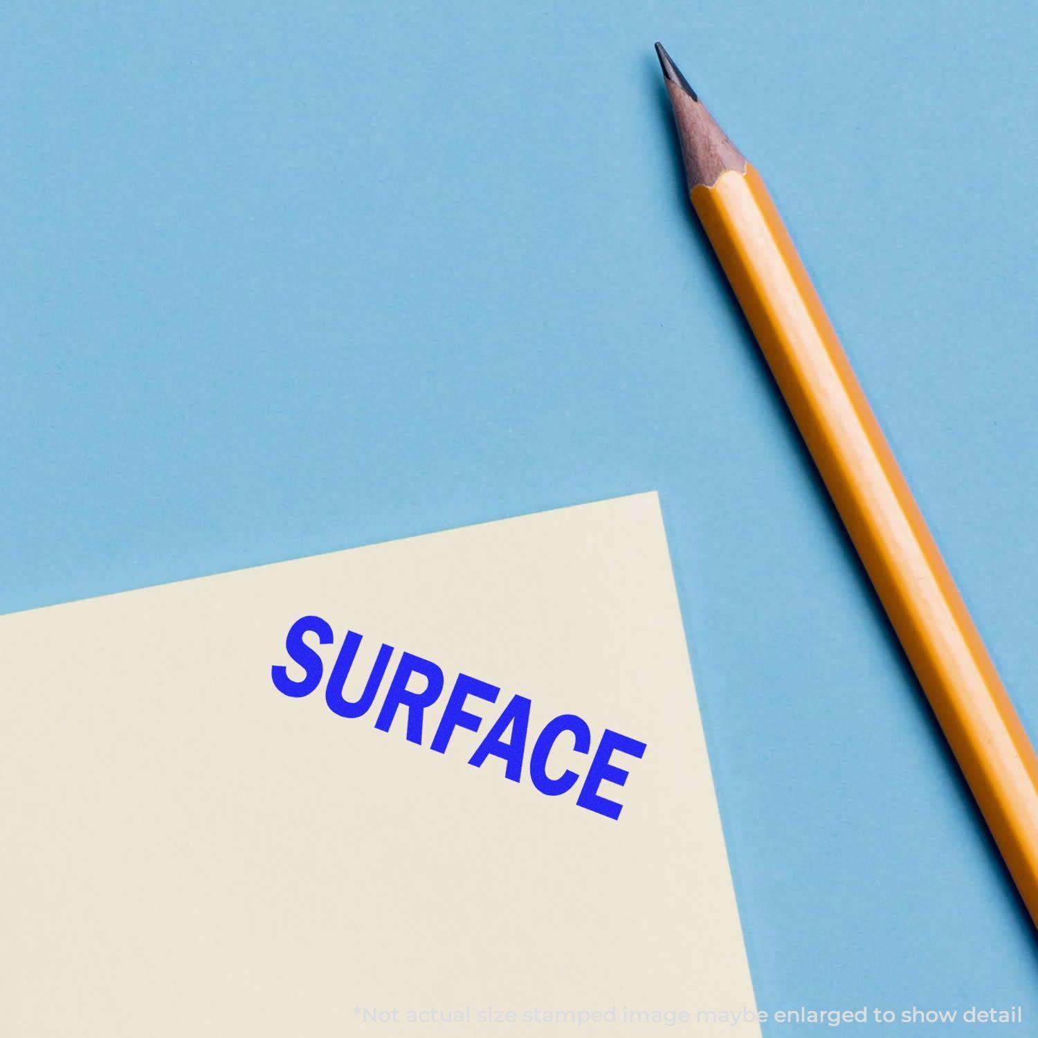 Yellow pencil next to a paper stamped with the word 'SURFACE' in blue ink using a Slim Pre-Inked Surface Stamp on a blue background.