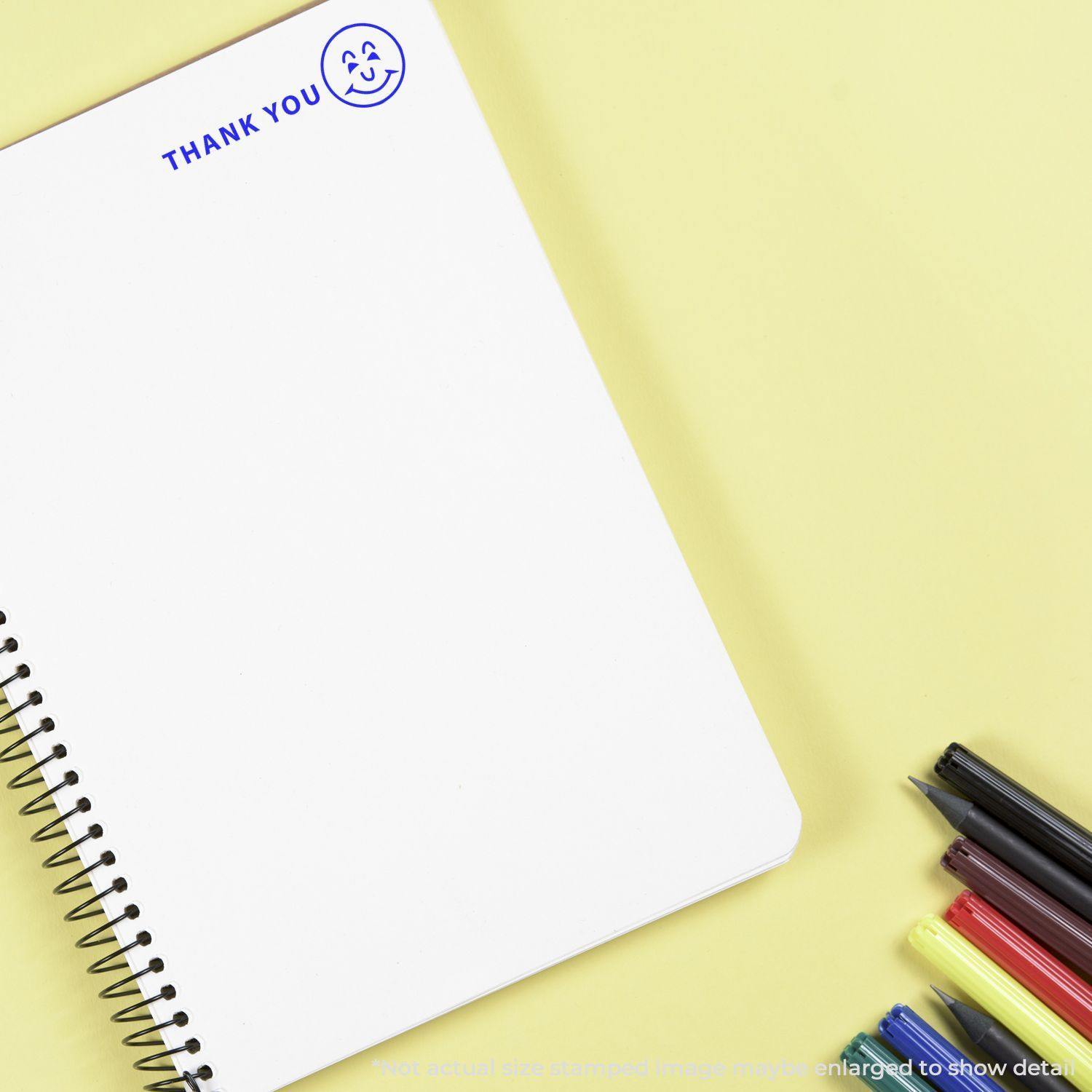 Large Self Inking Thank You with Smiley Stamp on a white notebook, with colorful pens nearby on a yellow background.