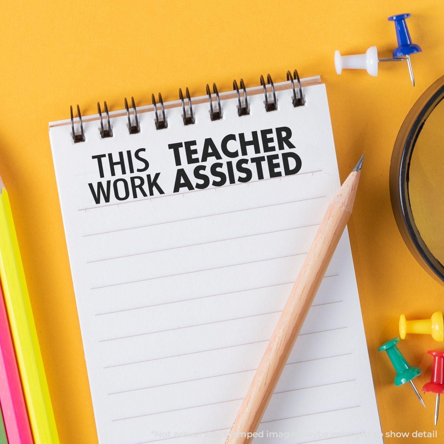 Large Pre-Inked This Work Teacher Assisted Stamp on a notepad with a pencil, colorful pins, and highlighters on a yellow background.