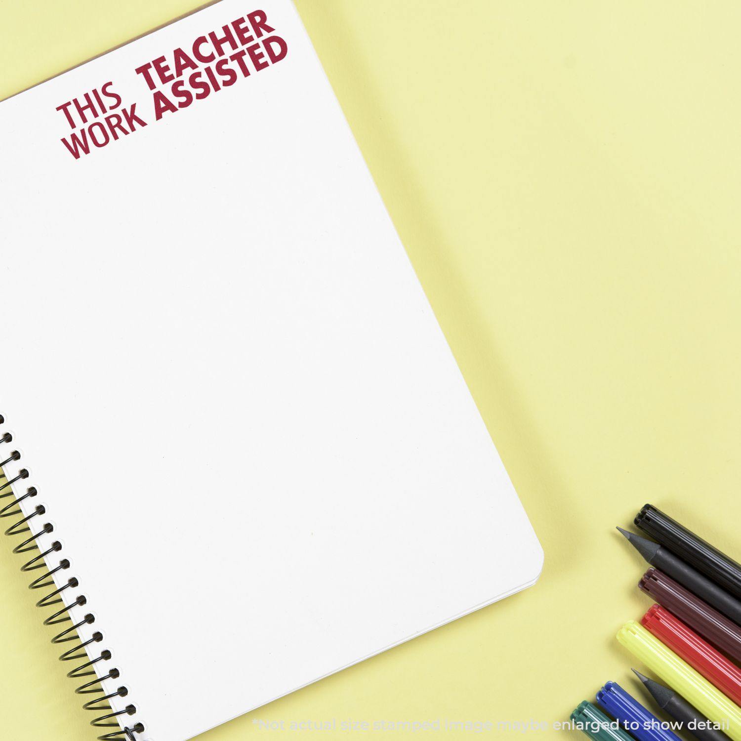 Large Self Inking This Work Teacher Assisted Stamp used on a notebook, with colored pens placed beside it on a yellow background.