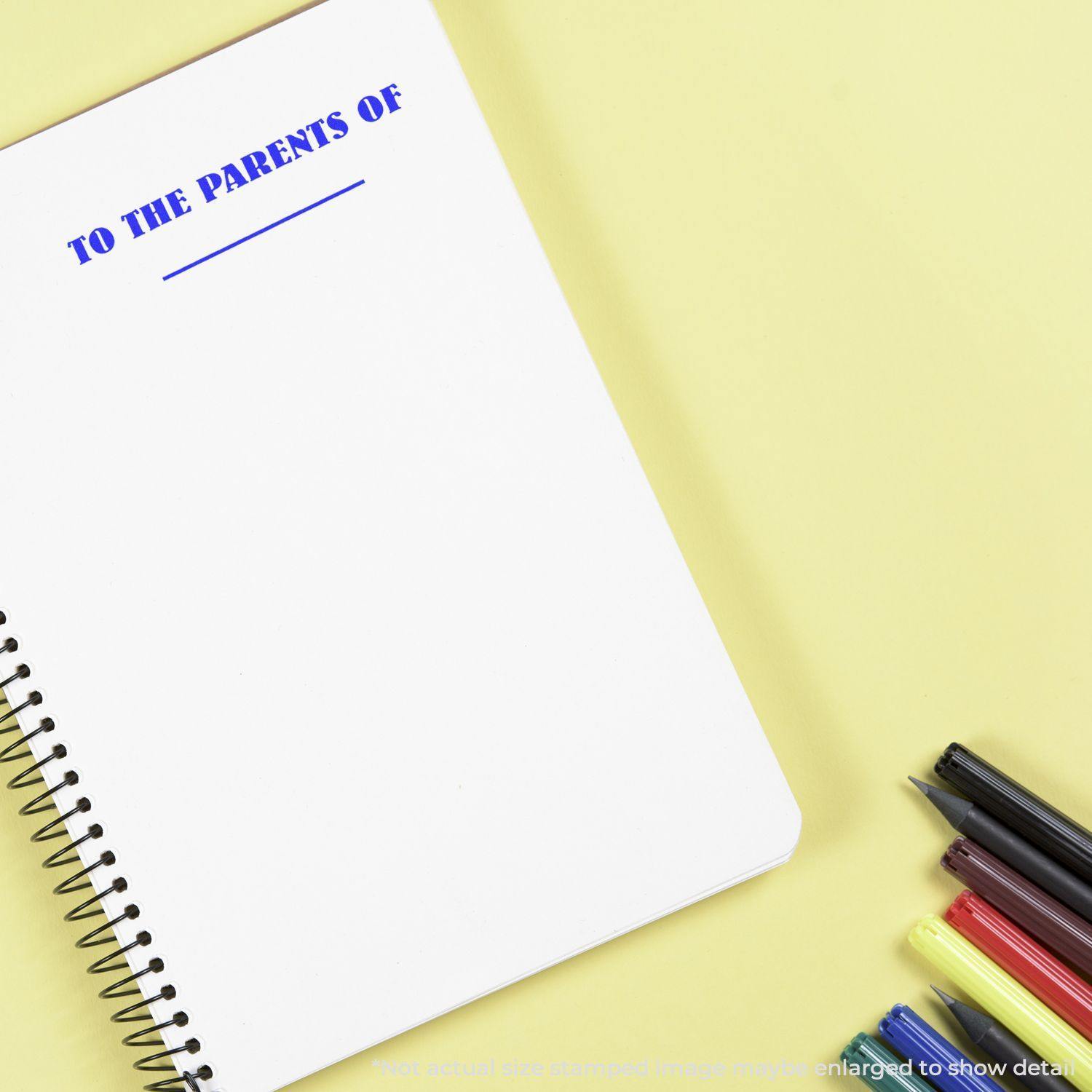 Notebook stamped with 'TO THE PARENTS OF' using a Slim Pre-Inked To The Parents Of Stamp, surrounded by colorful pens on a yellow background.