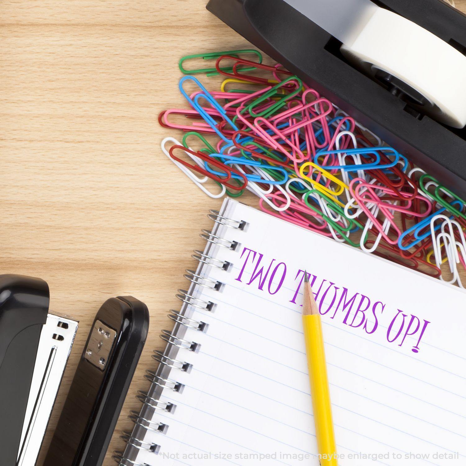 Large Pre-Inked Two Thumbs Up Stamp used on a notebook with a pencil, stapler, tape dispenser, and colorful paperclips on a wooden desk.