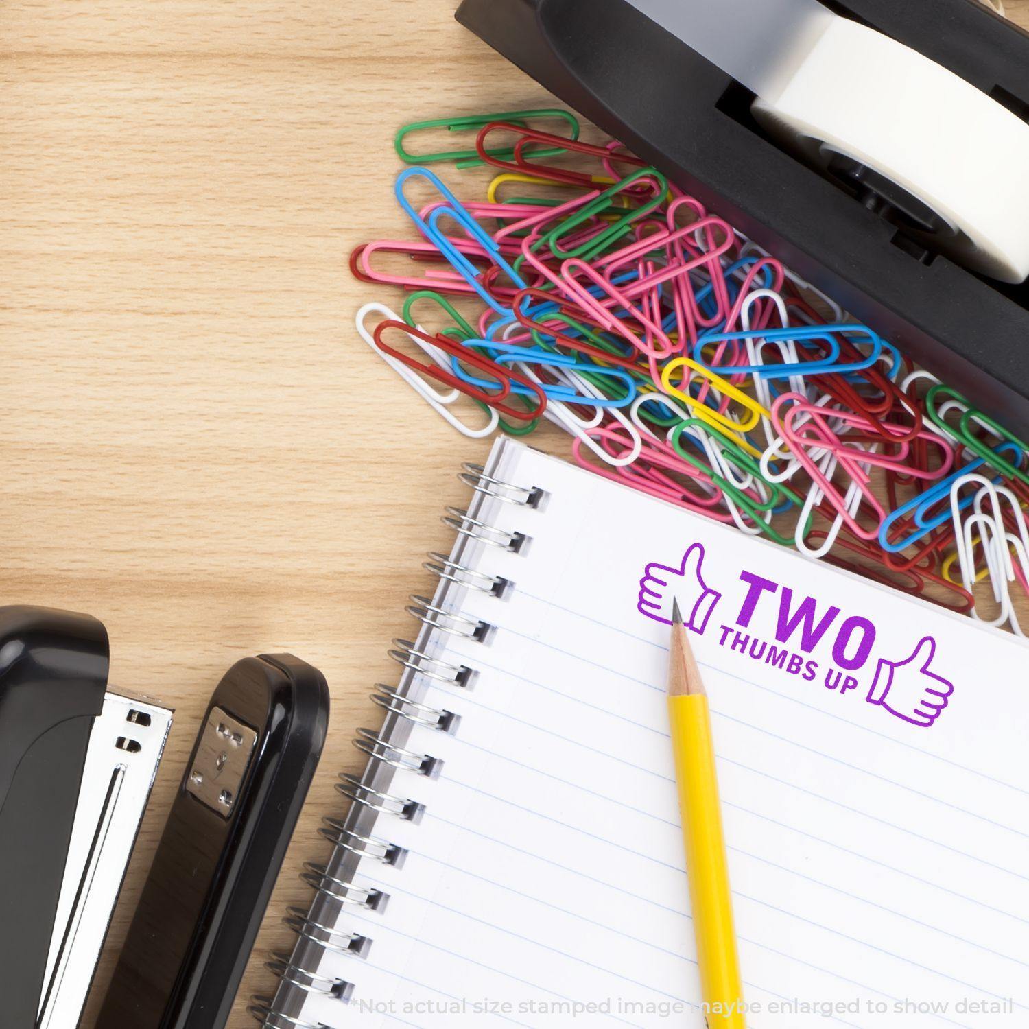 Two Thumbs Up with Thumb Icon Rubber Stamp on a notebook, surrounded by colorful paperclips, a stapler, and a tape dispenser.