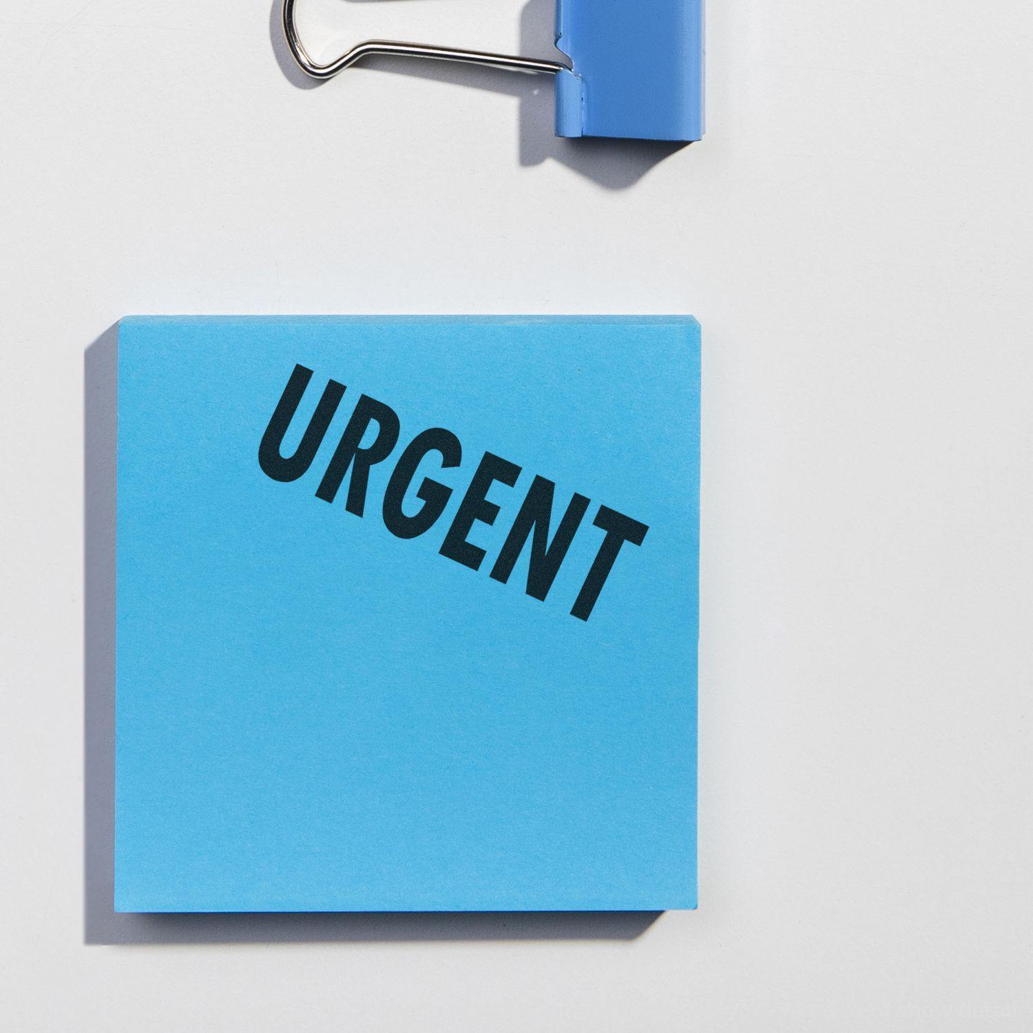 Blue sticky note with URGENT stamped in bold black letters, next to a blue binder clip on a white background.