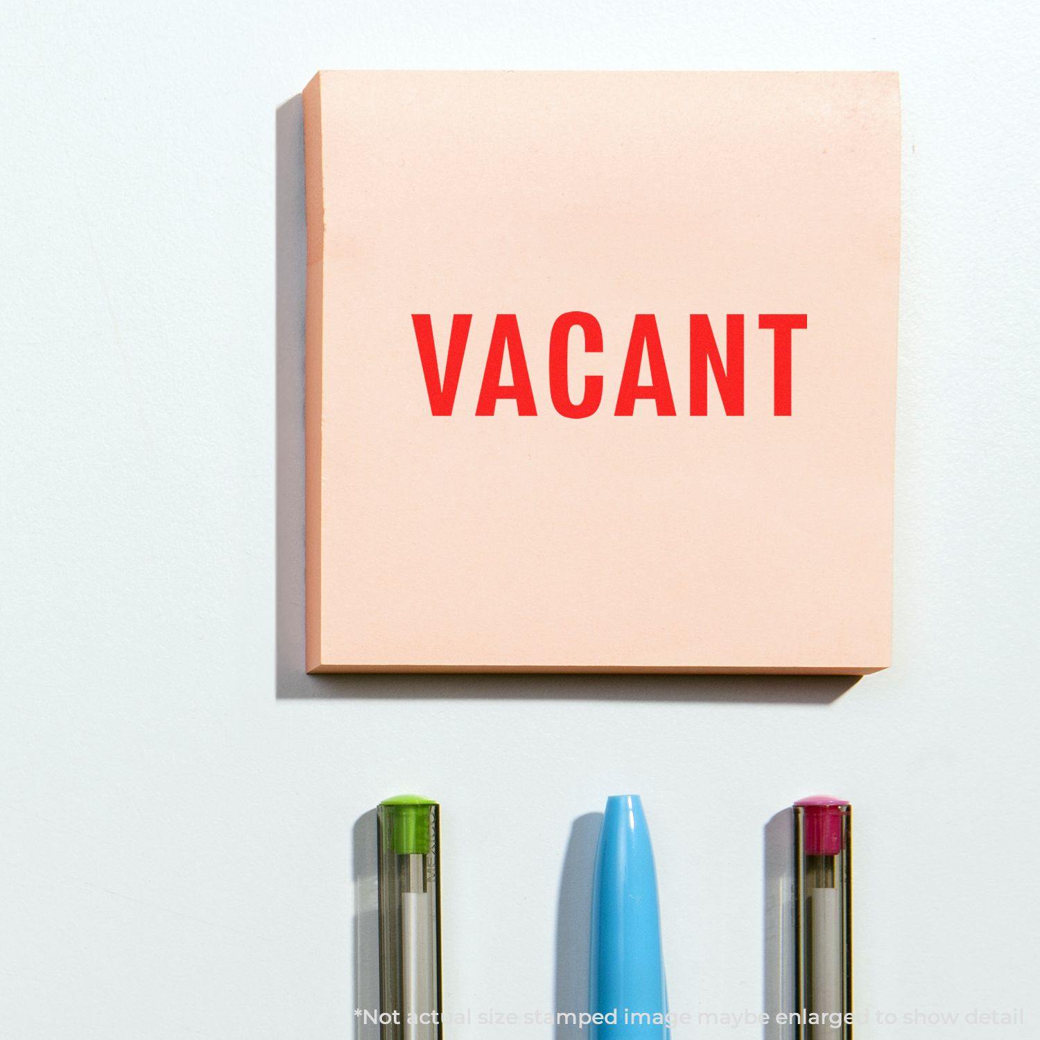 Vacant rubber stamp impression on a pink sticky note, placed above three pens with different colored tips on a white surface.