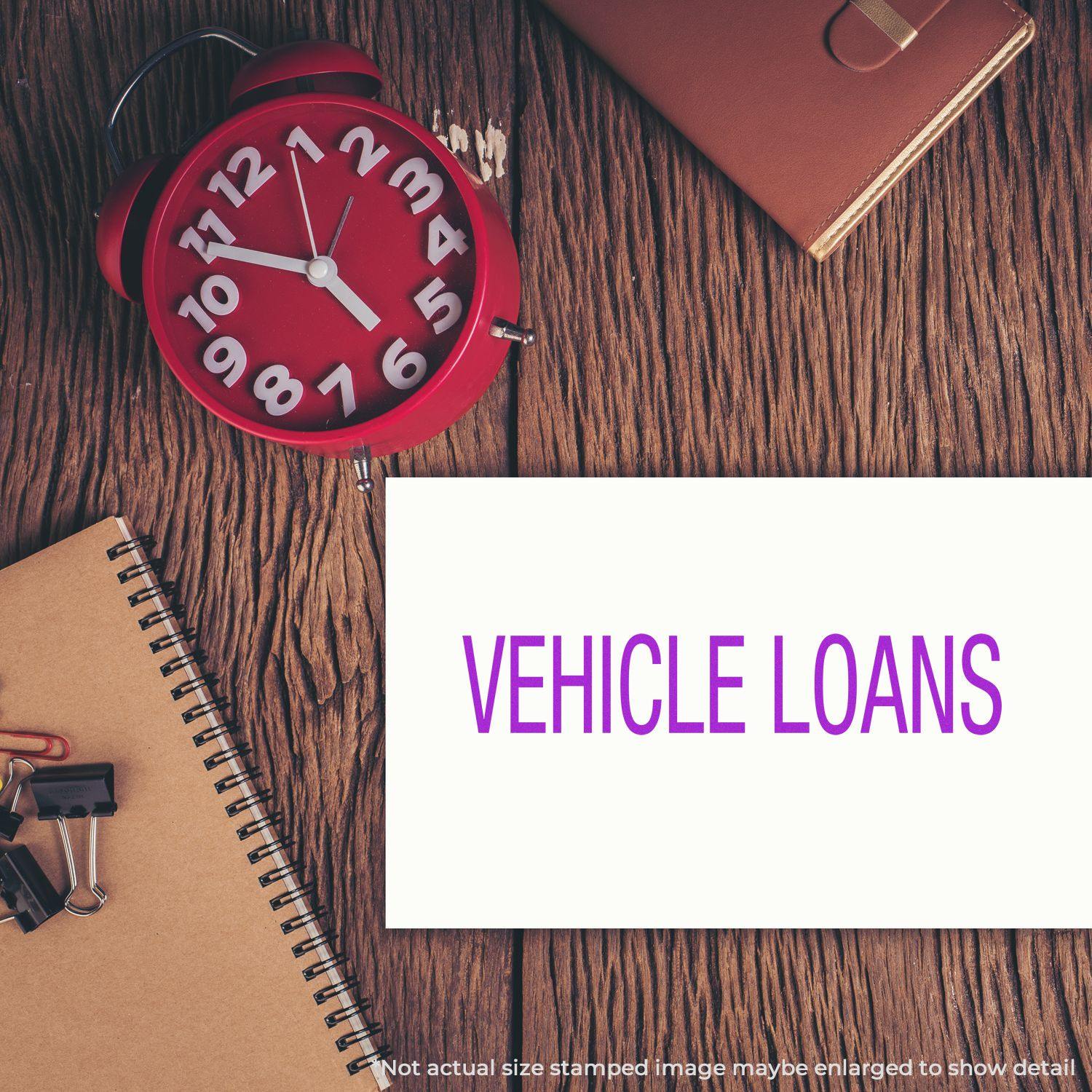 A red clock, notebook, and office supplies on a wooden desk with a stamped paper reading VEHICLE LOANS using the Self Inking Vehicle Loans Stamp.