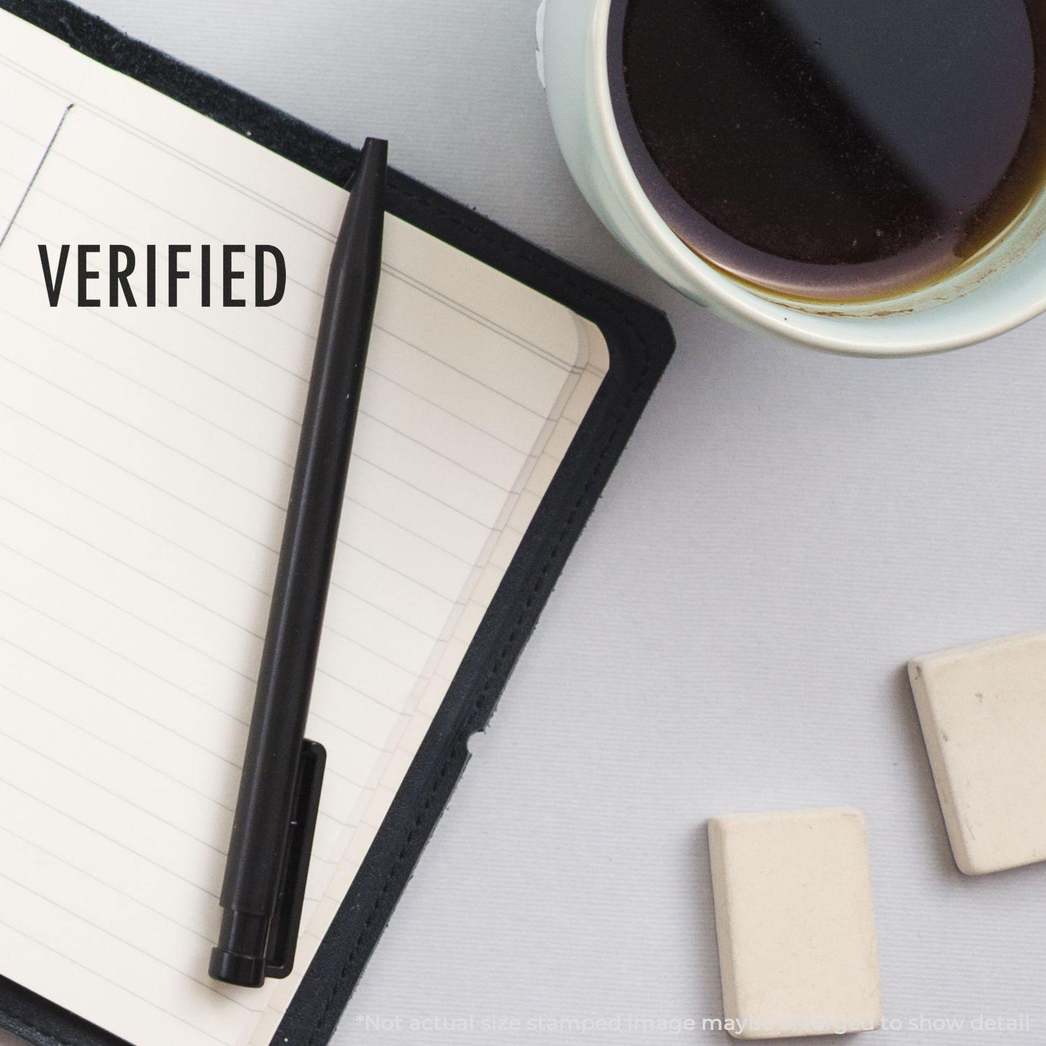 Large Self Inking Verified Stamp used on a notebook, with a pen, erasers, and a cup of coffee nearby.