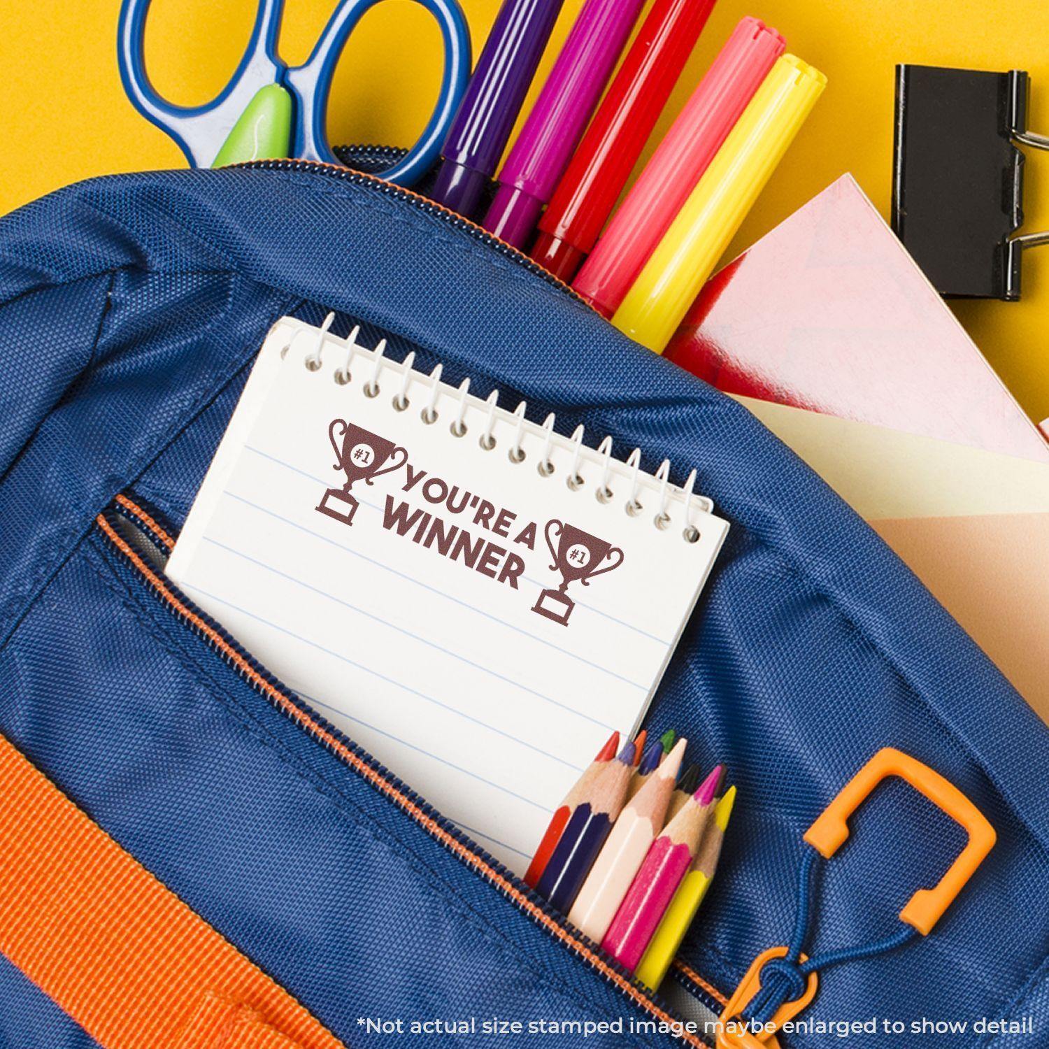 Slim Pre-Inked You're a Winner Stamp on a notepad in a blue backpack with colored pencils, markers, scissors, and paper clips.