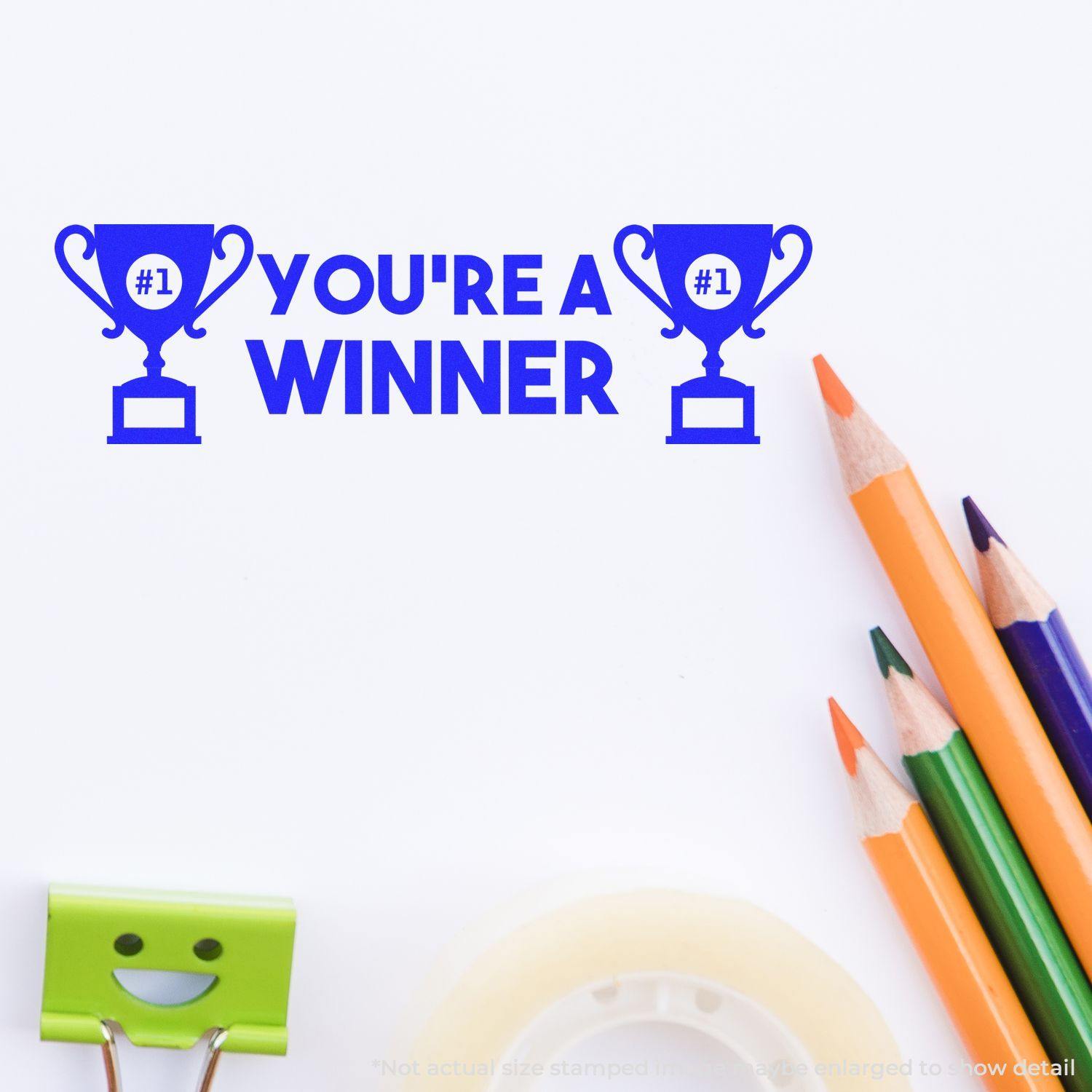 You're a Winner Rubber Stamp in blue ink with trophy icons, surrounded by colorful pencils, a smiley clip, and tape on a white surface.