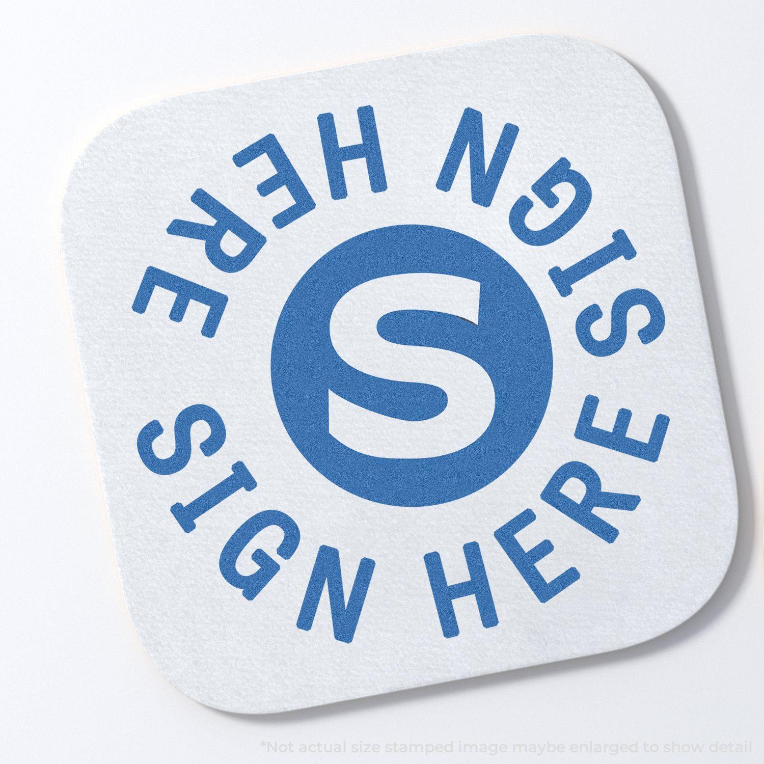 Round Sign Here Xstamper Stamp imprint on a white square paper with rounded corners, featuring a blue S in the center and SIGN HERE text.