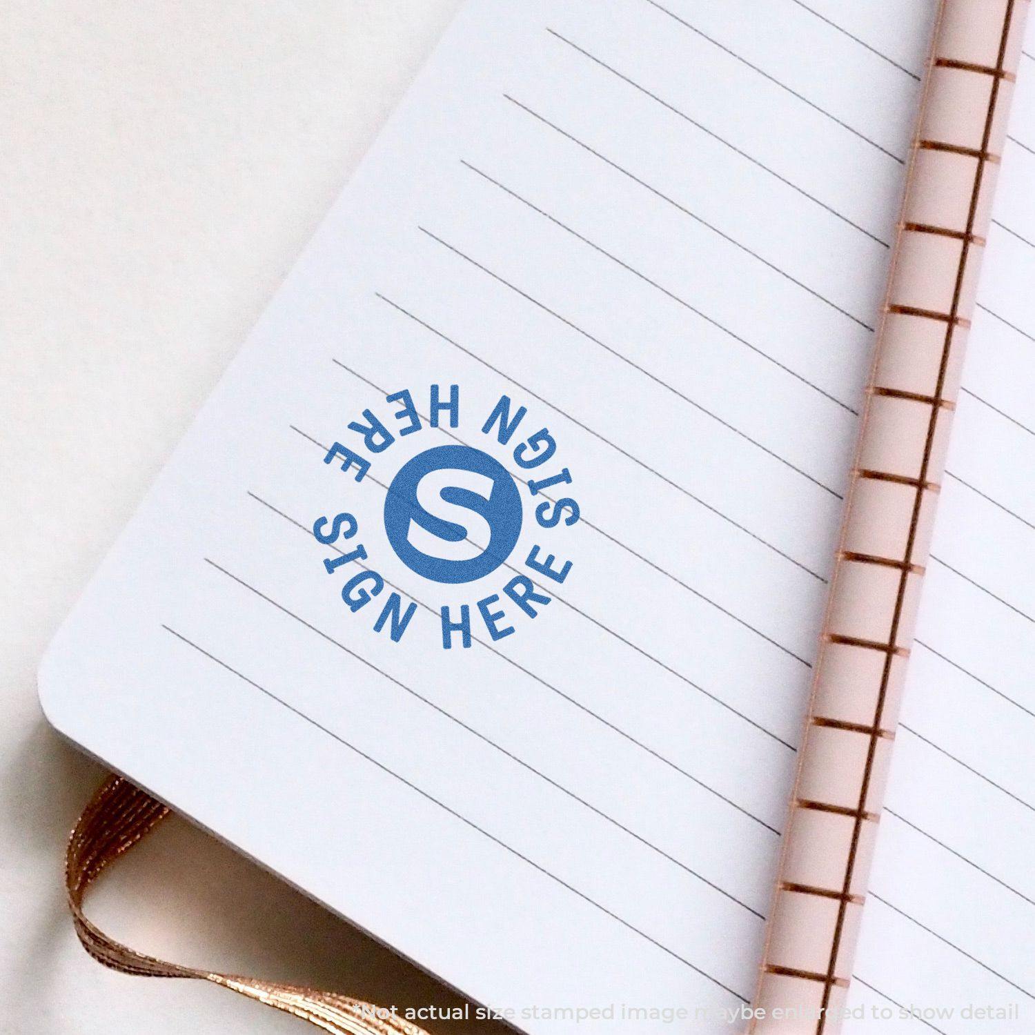 Round Sign Here Xstamper Stamp in blue ink on a lined notebook page with a copper spiral binding.